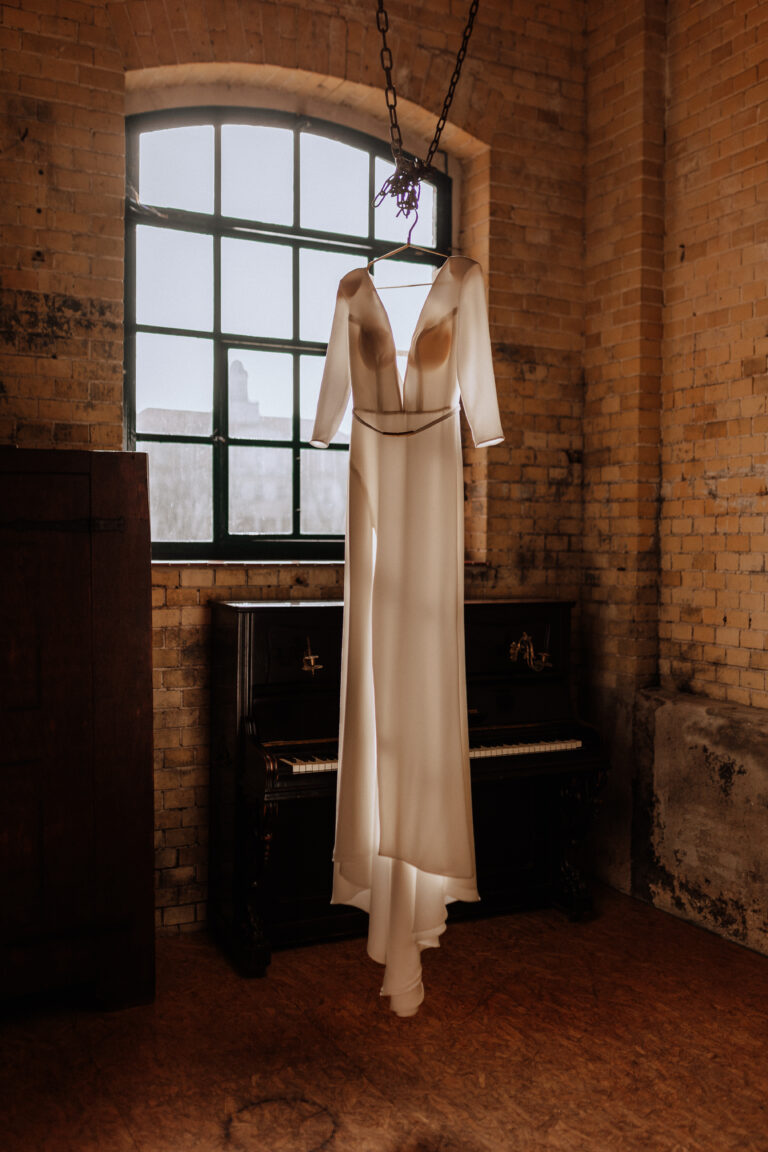 Ein langärmeliges weißes Kleid hängt vor einem Fenster über einem Klavier in einem gemauerten Raum, akzentuiert mit eleganter Hochzeitsdeko - perfekt für eine romantische Brandenburger Feier.