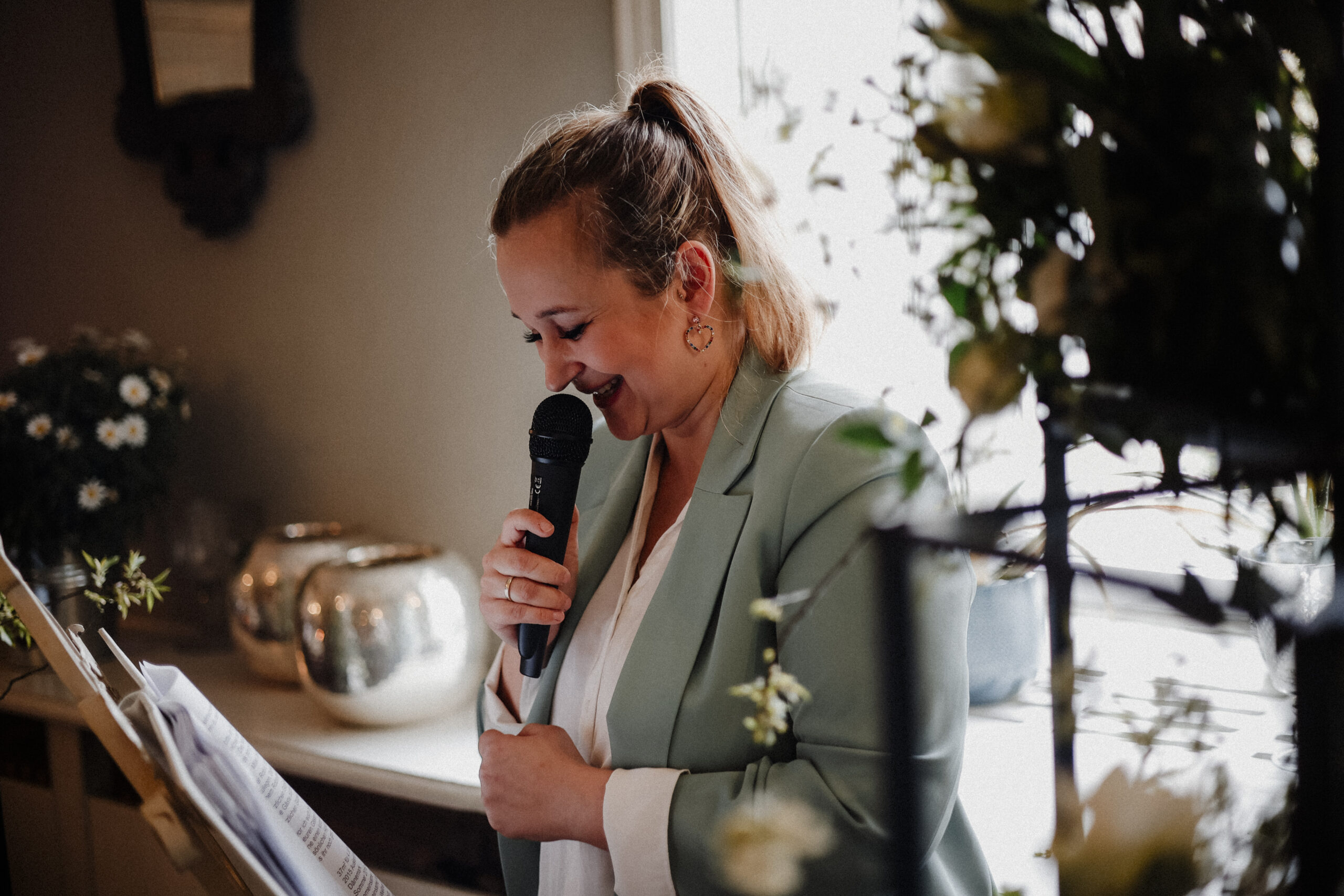 Eine Frau in einem mintfarbenen Blazer lächelt, während sie in ein Mikrofon spricht. Sie steht in einem Raum in der Nähe von Blumen und Papieren - die perfekte Inspiration für jeden Hochzeitsplaner in Brandenburg, der nach eleganten Hochzeitsdeko-Ideen sucht.