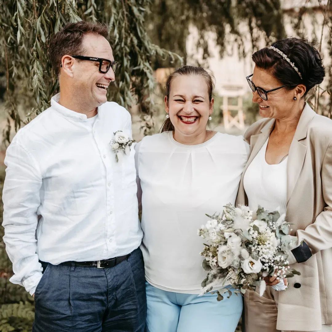 Drei Erwachsene lächeln gemeinsam im Freien in Brandenburg; einer hält einen Blumenstrauß, alle sind leicht und halbförmig gekleidet - perfekt für eine Zeremonie der Freien Traurednerin bei Berlin.