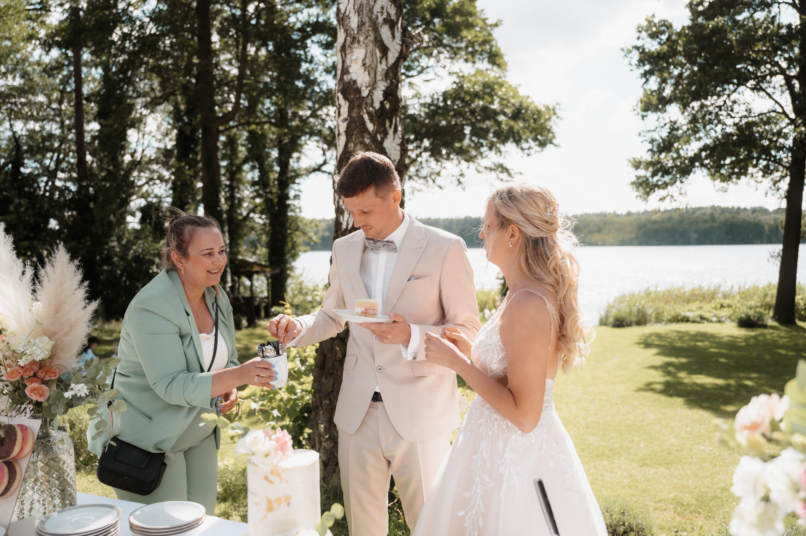 Drei Personen, darunter Braut und Bräutigam, stehen an einem sonnigen Tag im Freien an einer Kuchentafel in der Nähe eines Sees und bilden die perfekte Kulisse für einen Hochzeitsplaner in Brandenburg.