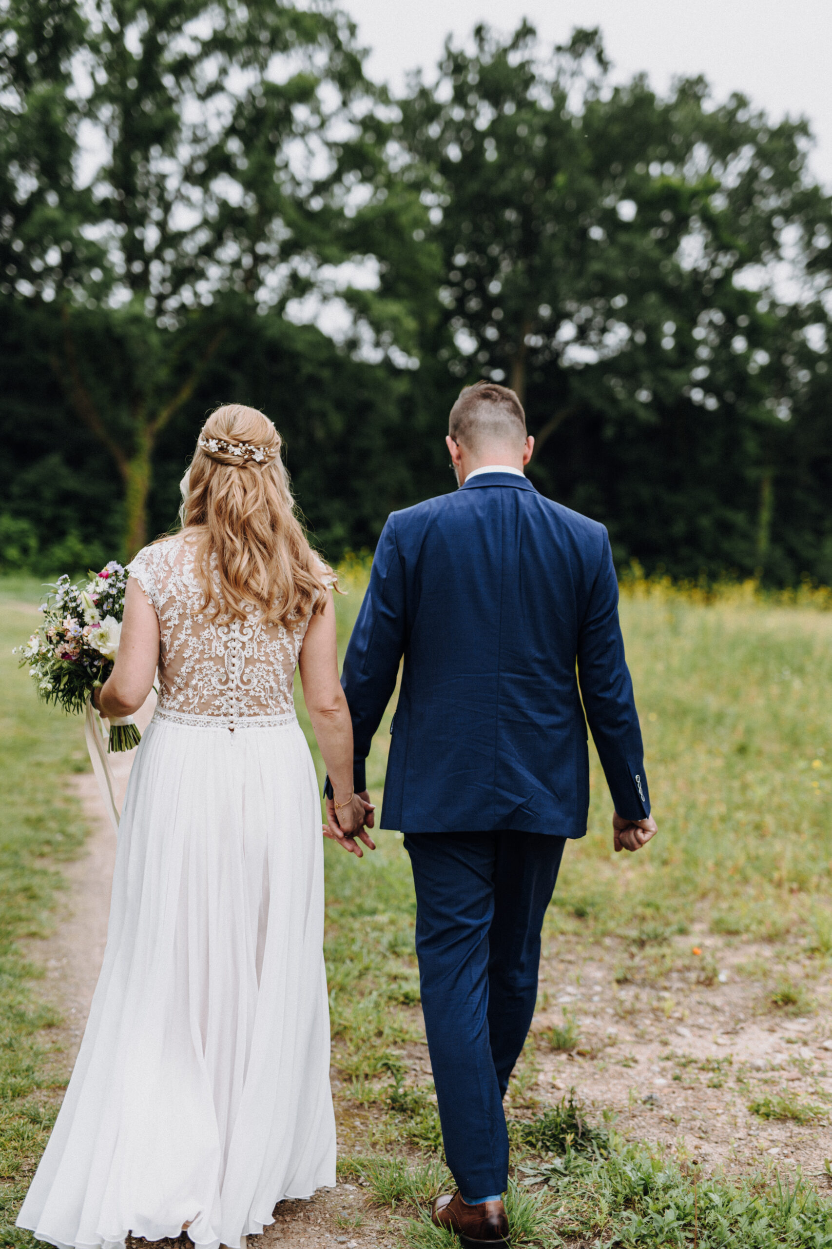 Eine Braut und ein Bräutigam gehen Hand in Hand über einen grasbewachsenen Weg, mit dem Rücken zur Kamera - ein unvergesslicher Moment, perfekt eingefangen für jeden Hochzeitsplaner oder jede Freie Traurednerin in Berlin, die Inspiration suchen.