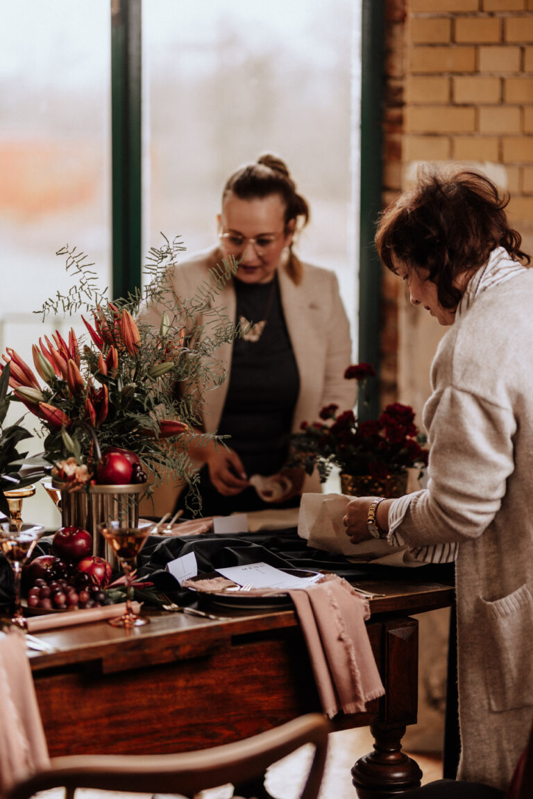 Zwei Frauen, möglicherweise eine Freie Traurednerin oder Hochzeitsplanerin, arrangieren Blumen und Papiere auf einem Holztisch in einem Haus in Brandenburg, umgeben von Vasen mit roten Lilien und Grünzeug.