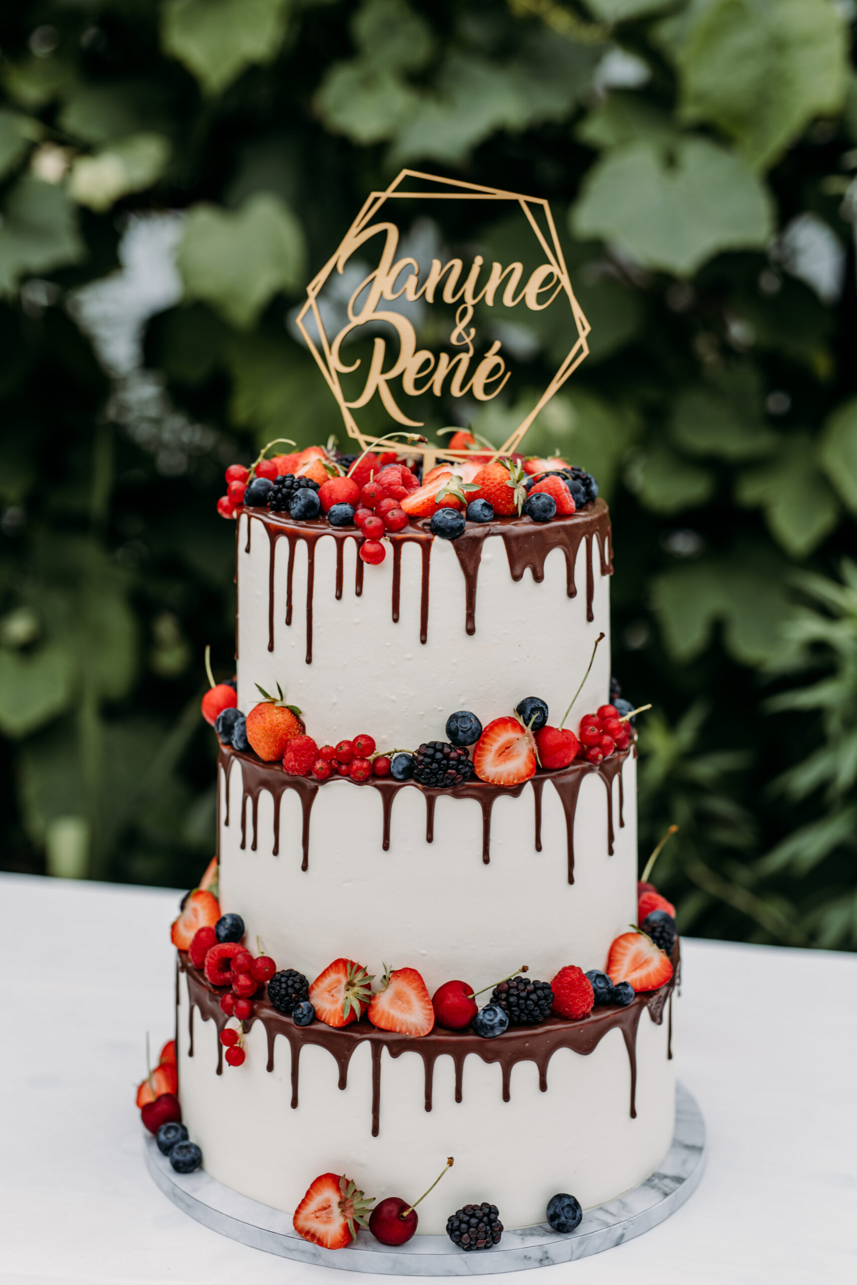 Dreistöckige weiße Torte mit Schokotropfen, gekrönt mit Beeren und einem "Janine & René"-Schild - die perfekte Hochzeitsdeko für eine elegante Feier im Freien in Berlin oder Brandenburg.