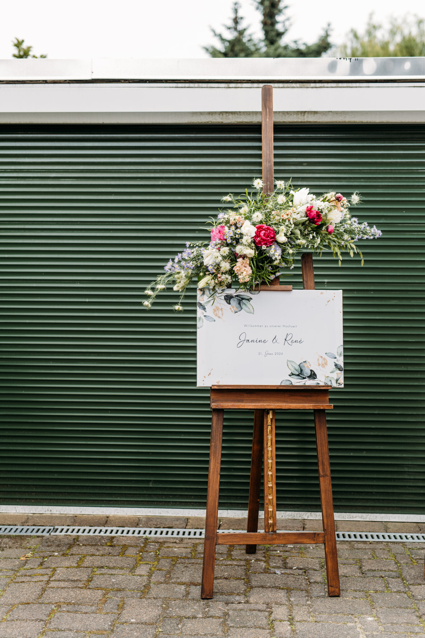 Florales Willkommensschild auf einer Holzstaffelei vor einem grünen Garagentor - die perfekte Inspiration für jeden Hochzeitsplaner oder jede Freie Traurednerin in Brandenburg.