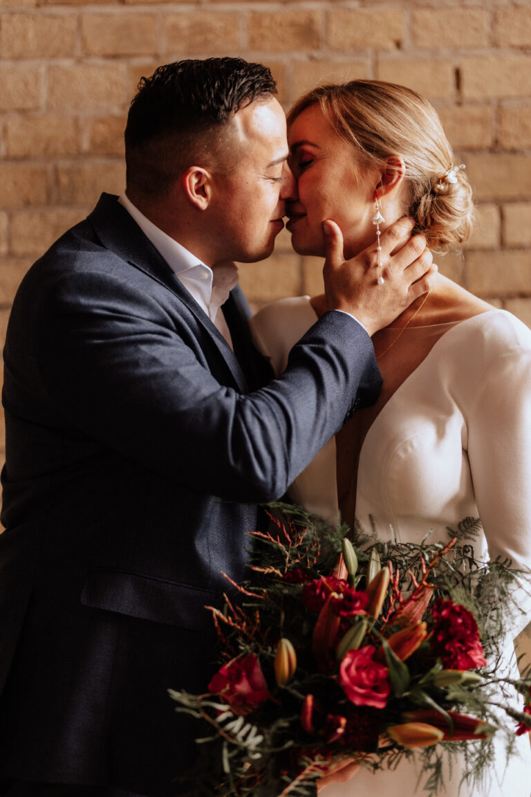 Ein Bräutigam hält sanft das Gesicht seiner Braut, während sie einen intimen Moment miteinander verbringen, umgeben von exquisiter Hochzeitsdeko und mit einem Strauß roter Blumen.
