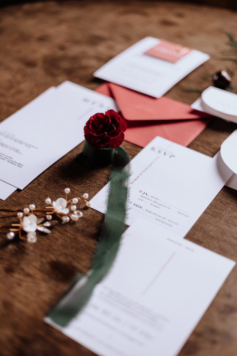 Flaches Layout einer eleganten Hochzeitsdeko: Hochzeitspapier, eine rote Blume und ein grünes Band auf einem Holztisch - die perfekte Inspiration für Feierlichkeiten in Berlin oder Brandenburg.