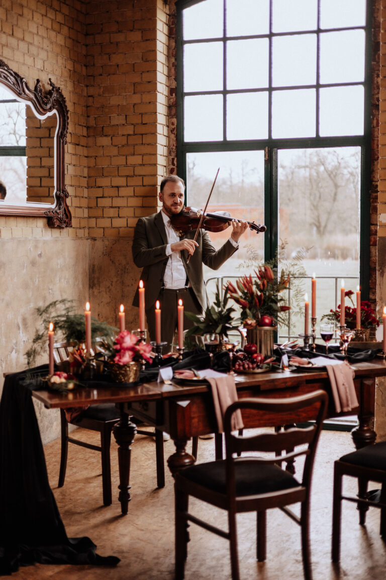 Ein Mann spielt Geige neben einem elegant gedeckten Esstisch mit Kerzen und Blumenschmuck, der eine romantische Atmosphäre schafft, die perfekt für die Hochzeitsdeko auf einer Berliner Feier ist.