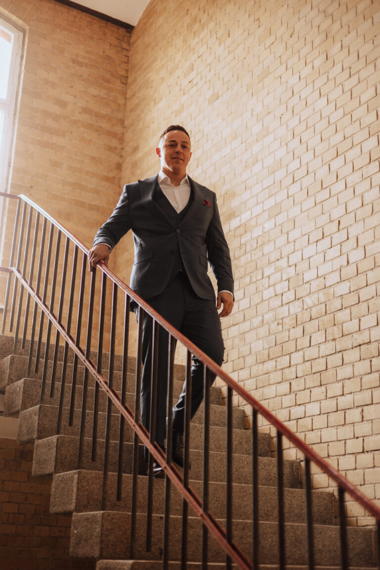 Ein Mann im Anzug steigt eine Treppe hinunter, die von Backsteinwänden gesäumt ist, und das Sonnenlicht, das durch ein nahe gelegenes Fenster fällt, erinnert an die Eleganz einer Veranstaltung der Brandenburger Hochzeitsplaner.