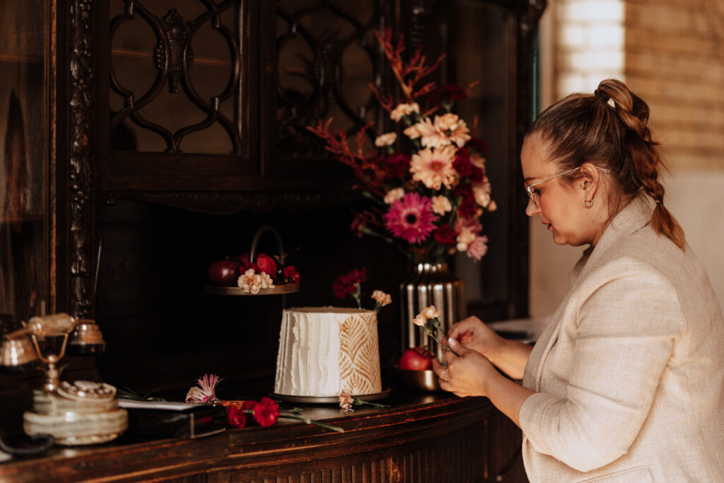 Eine Frau dekoriert eine weiße Torte mit Blumen auf einem Holztisch neben einer Vase mit farbenfrohen Blumen, um die perfekte Hochzeitsdeko für eine brandenburgische Feier zu schaffen.