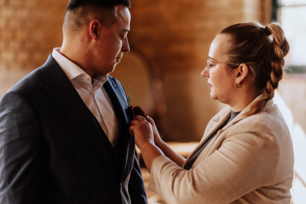 Eine Frau, vielleicht eine Hochzeitsplanerin, steckt einem Mann in einem warm beleuchteten Raum, umgeben von eleganter Hochzeitsdeko, eine Boutonnière an die Anzugsjacke.