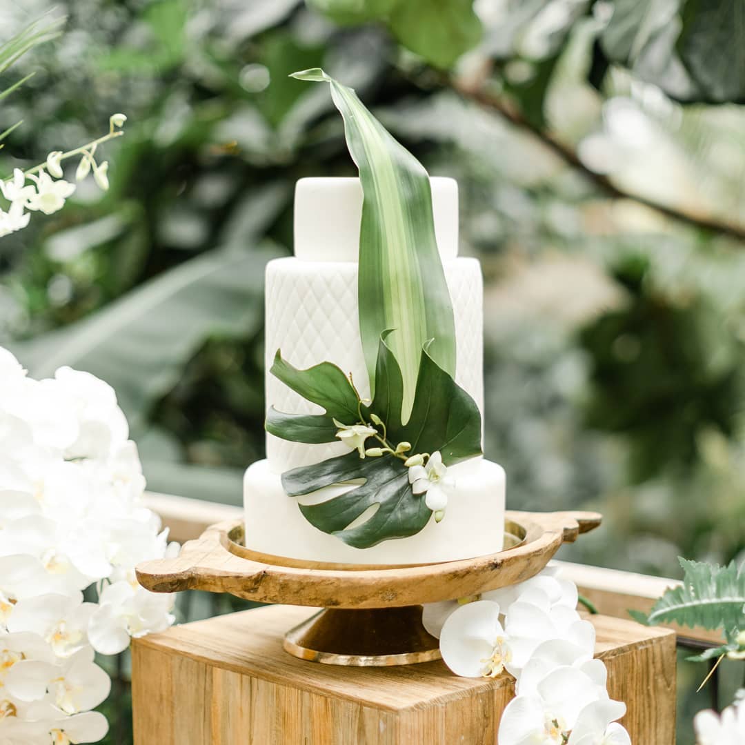 Weiße Stufentorte mit grünen tropischen Blättern und weißen Blumen auf einem Holzständer im Freien - perfekt für eine Berliner Hochzeit, die von einem Hochzeitsplaner geplant oder von einer Freien Traurednerin begleitet wird.