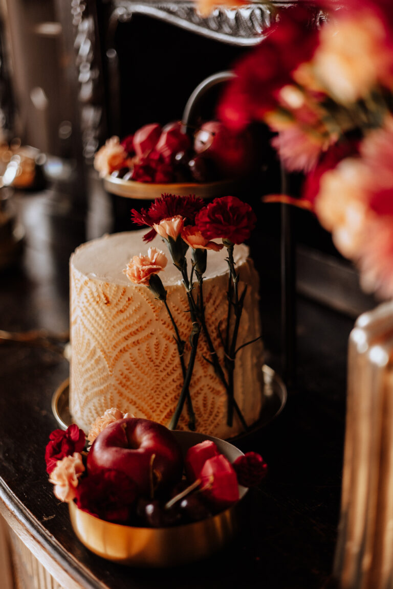 Hohe weiße Torte mit roter und rosa Blüte, inspiriert von der Hochzeitsdeko aus Berlin, neben roten Äpfeln in einer goldenen Schale.