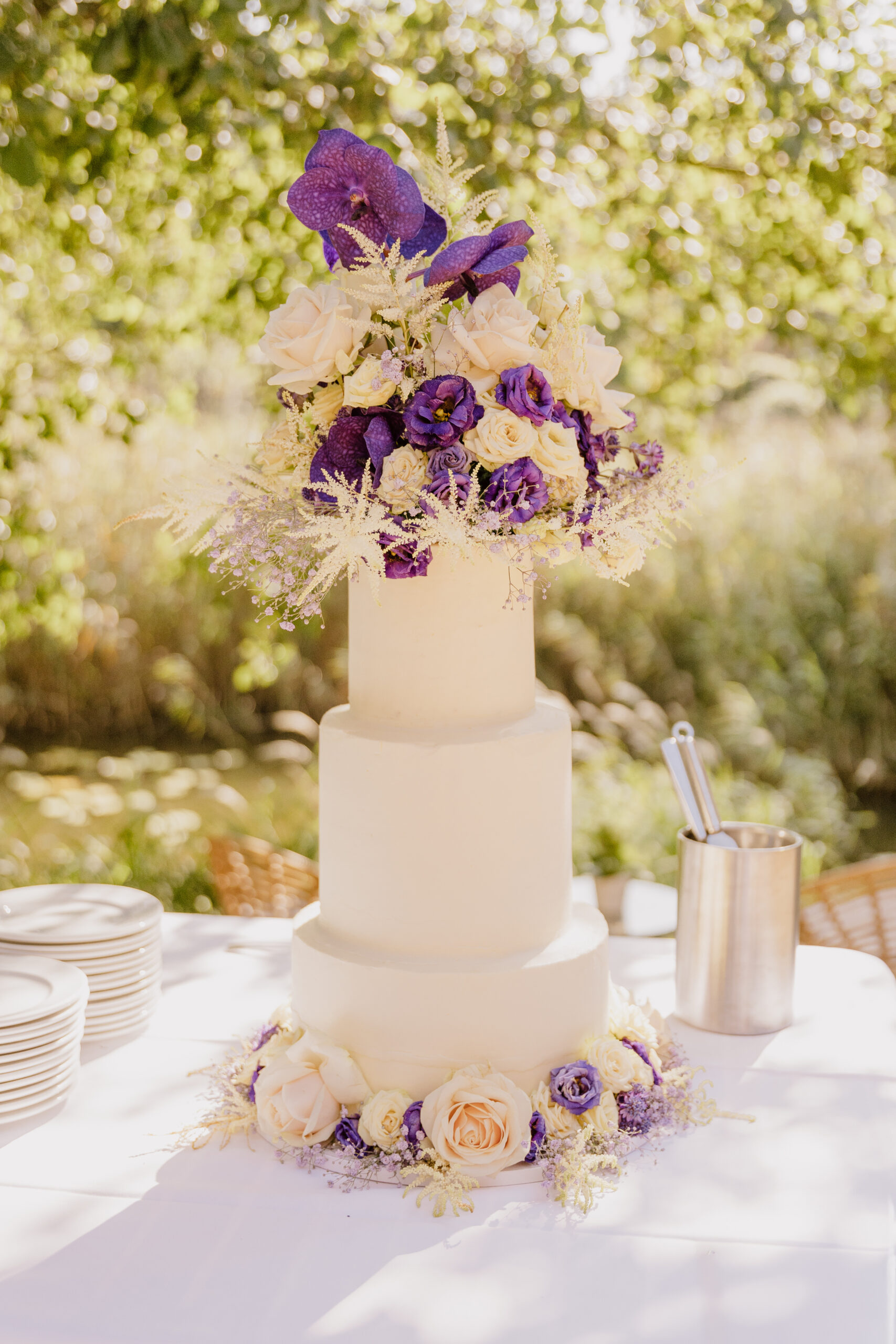 Dreistöckige weiße Hochzeitstorte mit lilafarbenen und cremefarbenen Blumen, die auf einem Tisch im Freien in Brandenburg steht.
