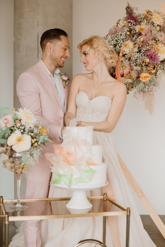 Braut und Bräutigam lächeln sich an, während sie eine weiße Hochzeitstorte anschneiden, umgeben von eleganter Hochzeitsdeko in Brandenburg, mit ihrer Freien Traurednerin in der Nähe.