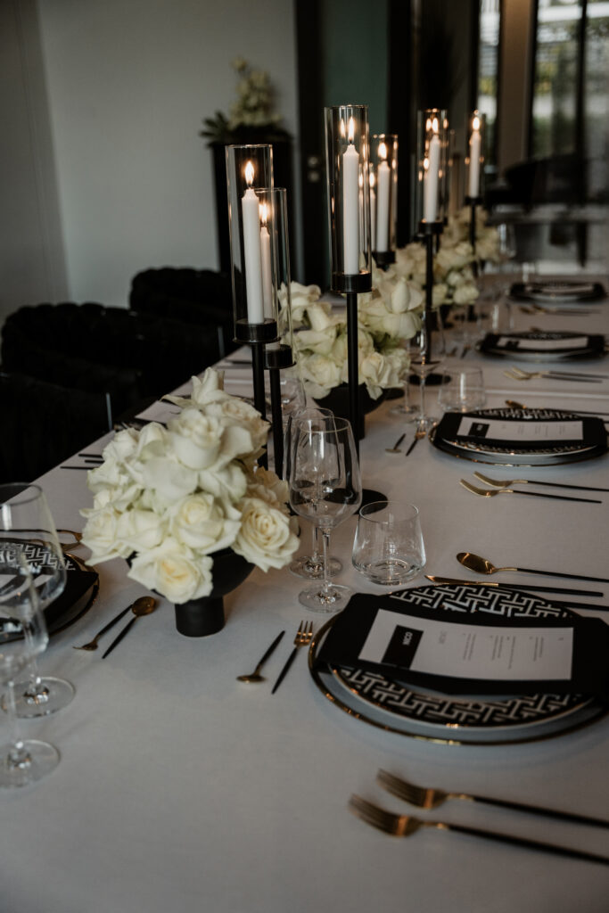 Elegant gedeckter Tisch mit weißen Rosen, schwarzen Kerzen, goldenem Besteck und schwarzen Tellern auf weißem Tischtuch - die perfekte Inspiration für einen Hochzeitsplaner oder eine Freie Traurednerin in Berlin.