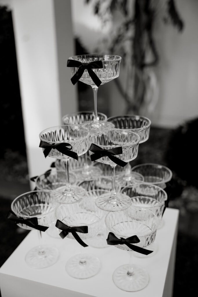 Eine Pyramide aus leeren Kristallgläsern, verziert mit kleinen schwarzen Schleifen, ausgestellt auf einem weißen Ständer - perfekt als Hochzeitsdeko für eine elegante Berliner Hochzeit.
