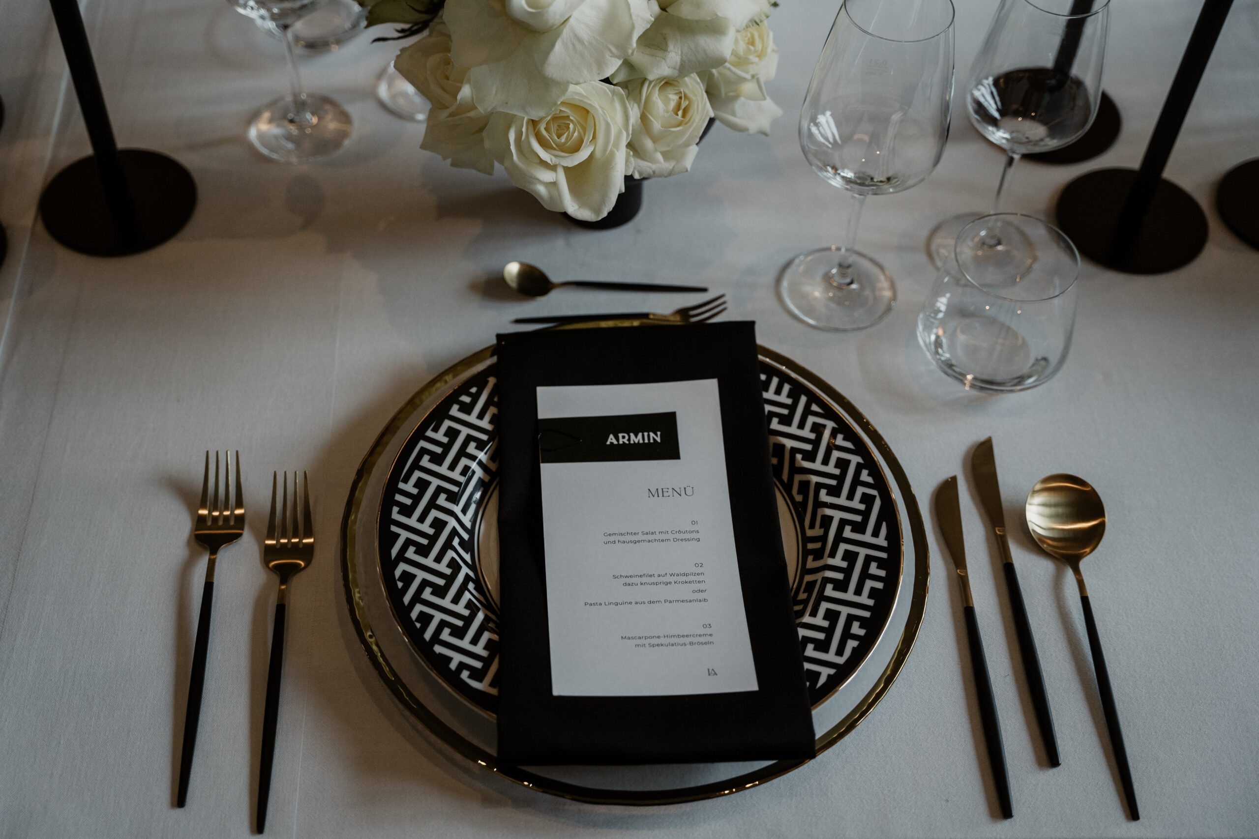 Elegant gedeckter Tisch mit schwarz-weißem Teller, Speisekarte, goldenem Besteck, Weingläsern und weißen Rosen - die perfekte Hochzeitsdeko für Ihre Feier in Brandenburg.