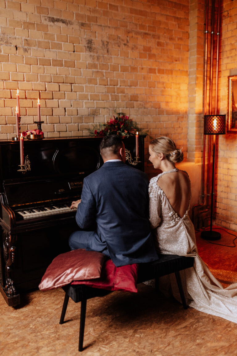 Braut und Bräutigam sitzen zusammen am Klavier in einem warm beleuchteten, rustikalen Raum mit Kerzen, Backsteinwänden und eleganter Hochzeitsdeko, die die romantische Atmosphäre noch verstärkt.