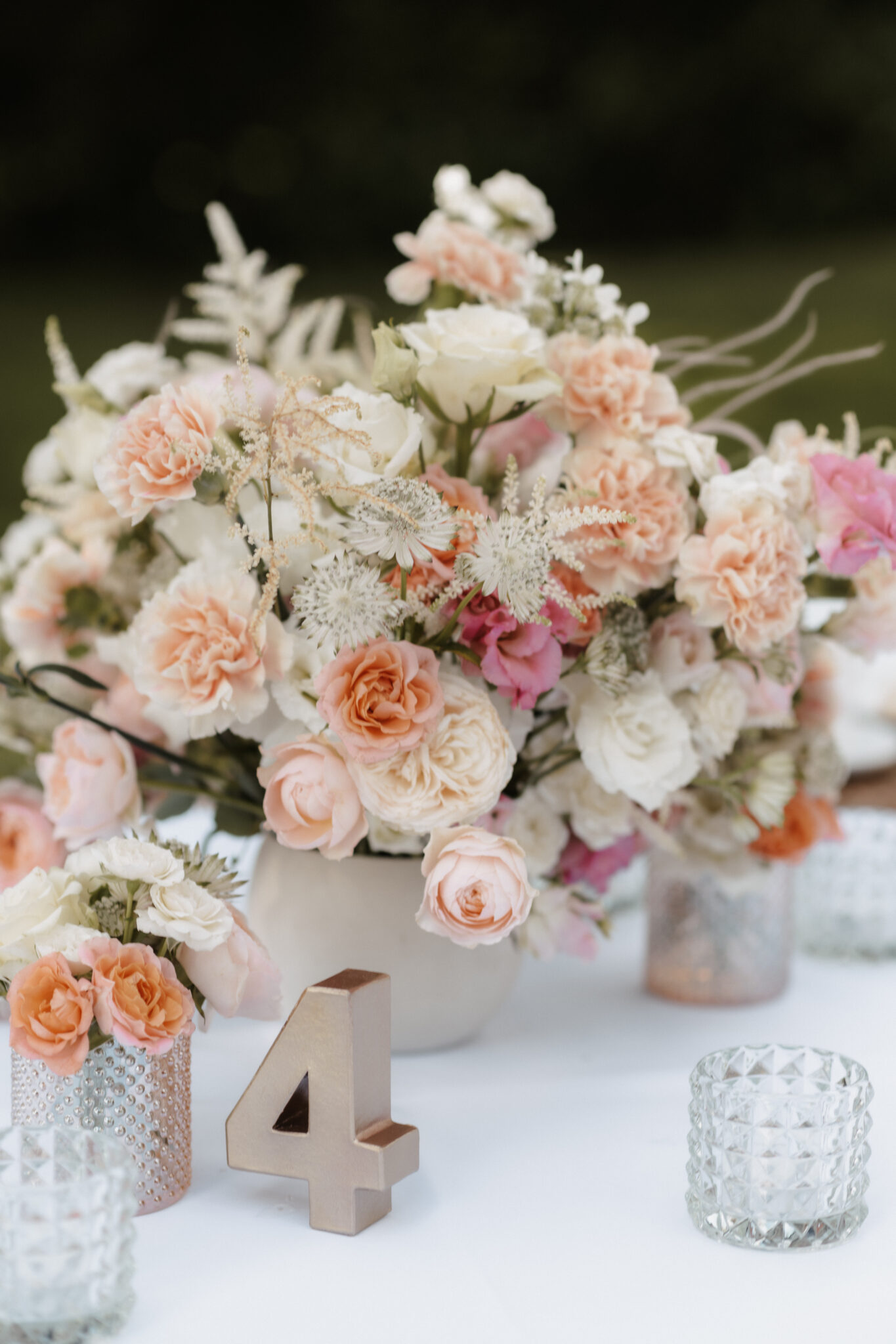 Ein florales Mittelstück mit rosa und weißen Blumen, Kerzenhaltern aus Glas und einer goldenen Tischnummer 4 - perfekt für eine Berliner Hochzeit oder für Paare, die mit einem Hochzeitsplaner in Brandenburg arbeiten.