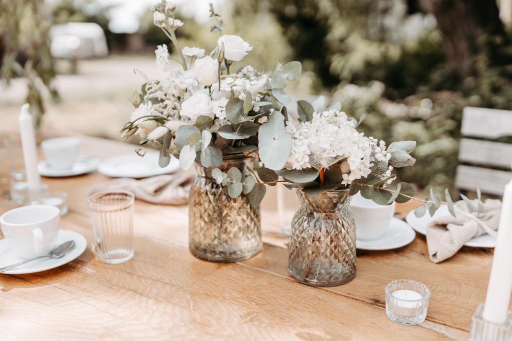 Blumenarrangements in Gläsern schmücken einen Holztisch, der für ein Essen im Freien in Brandenburg gedeckt ist - perfekt für einen Berliner Hochzeitsplaner, der nach Inspiration sucht. Teller und Tassen vervollständigen die charmante Szene.