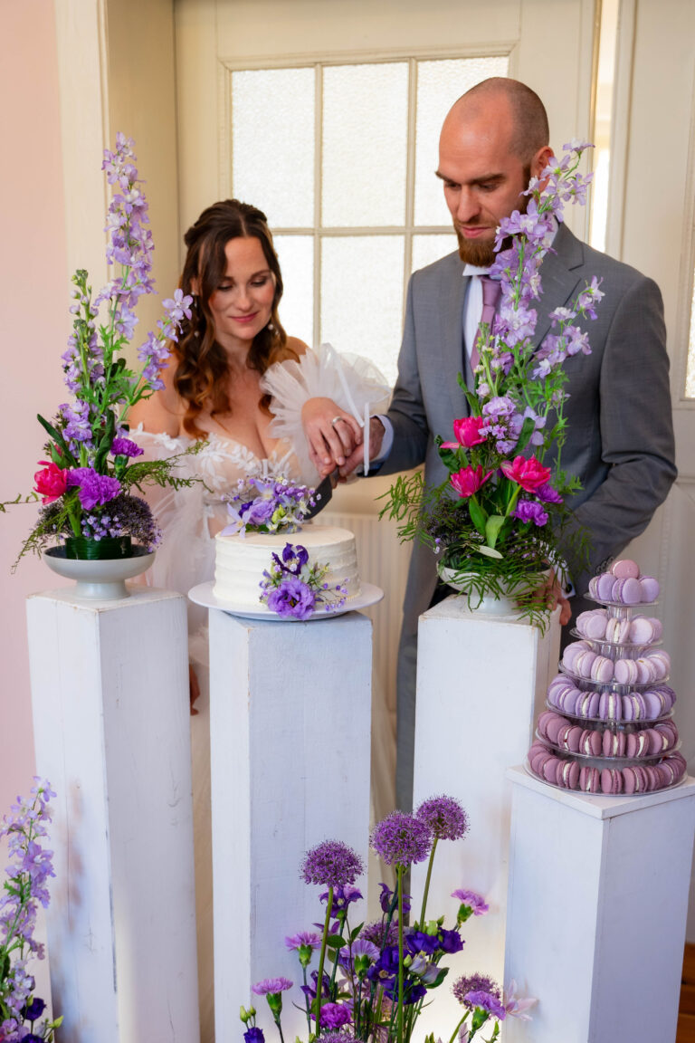 Braut und Bräutigam schneiden eine Torte an, die von lila Blumen und Macarons auf weißen Säulen umgeben ist - die perfekte Inspiration für jeden Hochzeitsplaner in Berlin oder Brandenburg.