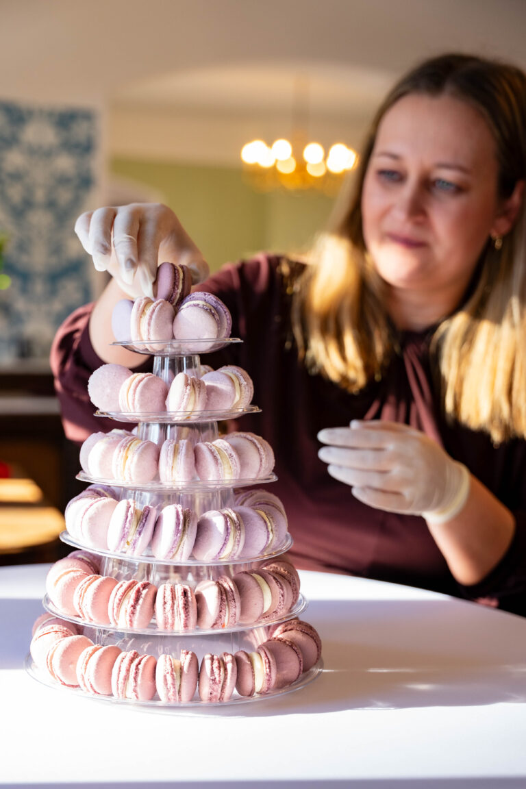 Eine Frau arrangiert rosafarbene Macarons auf einem gestuften Ständer und trägt dabei Handschuhe in einem gut beleuchteten Raum - die perfekte Hochzeitsdeko für jeden Hochzeitsplaner, der eine elegante Veranstaltung in Brandenburg vorbereitet.