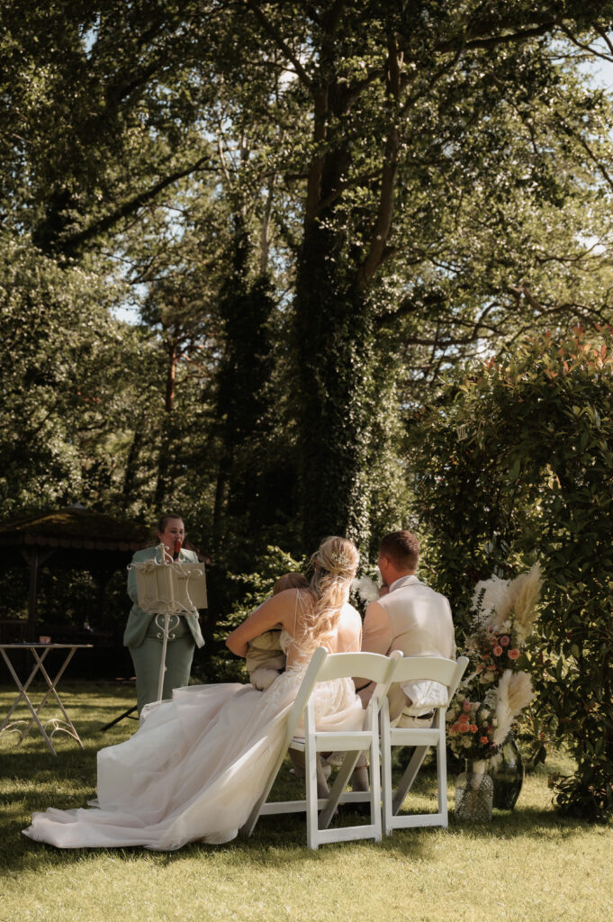 Braut und Bräutigam sitzen im Freien in Brandenburg und lauschen dem Trauredner bei einer sonnigen Gartenhochzeit mit eleganter Hochzeitsdeko.