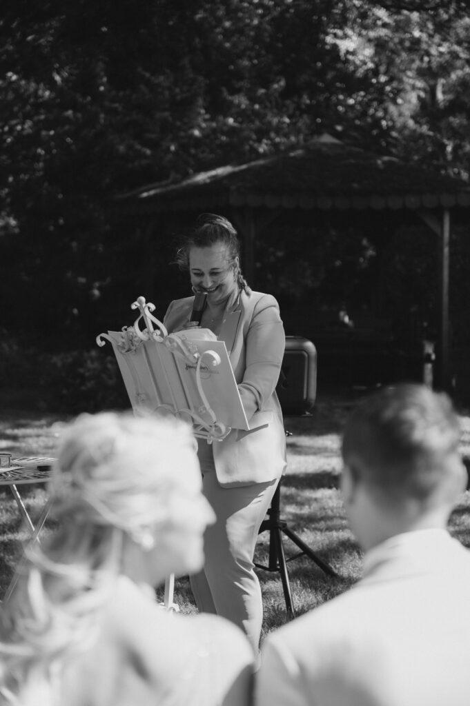 Eine Freie Traurednerin spricht bei einer Veranstaltung im Freien in Berlin und lächelt, während im Vordergrund zwei Personen zuhören.