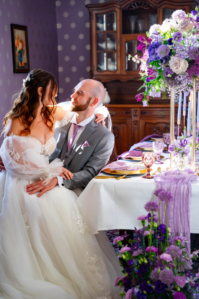 Die Braut sitzt auf dem Schoß des Bräutigams an einer Hochzeitstafel in Brandenburg, die mit lila Blumen und elegantem Geschirr geschmückt ist - die perfekte Inspiration für jeden Hochzeitsplaner in Berlin.