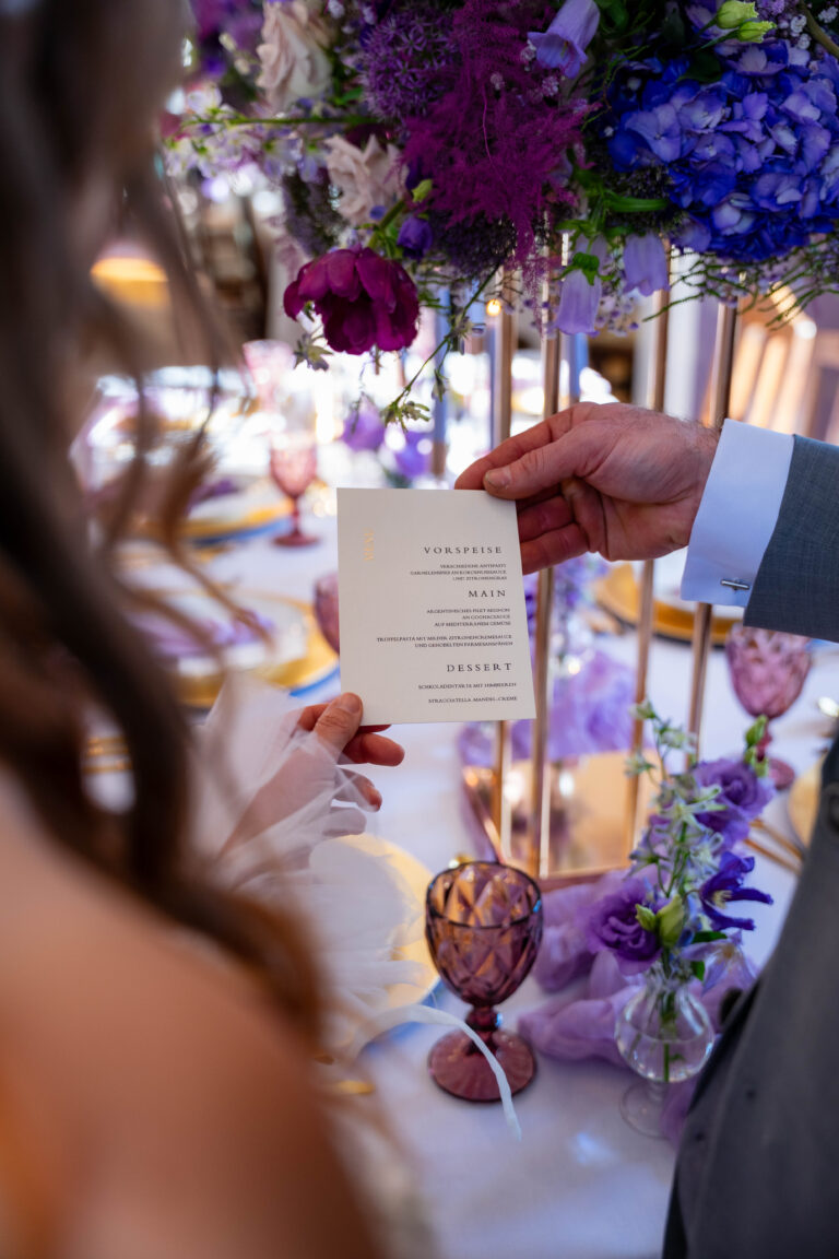 Eine Person hält eine Hochzeitsmenükarte an einem elegant dekorierten Tisch mit lila Blumen und Gläsern, die perfekte Inspiration für jeden Hochzeitsplaner in Berlin oder Brandenburg.