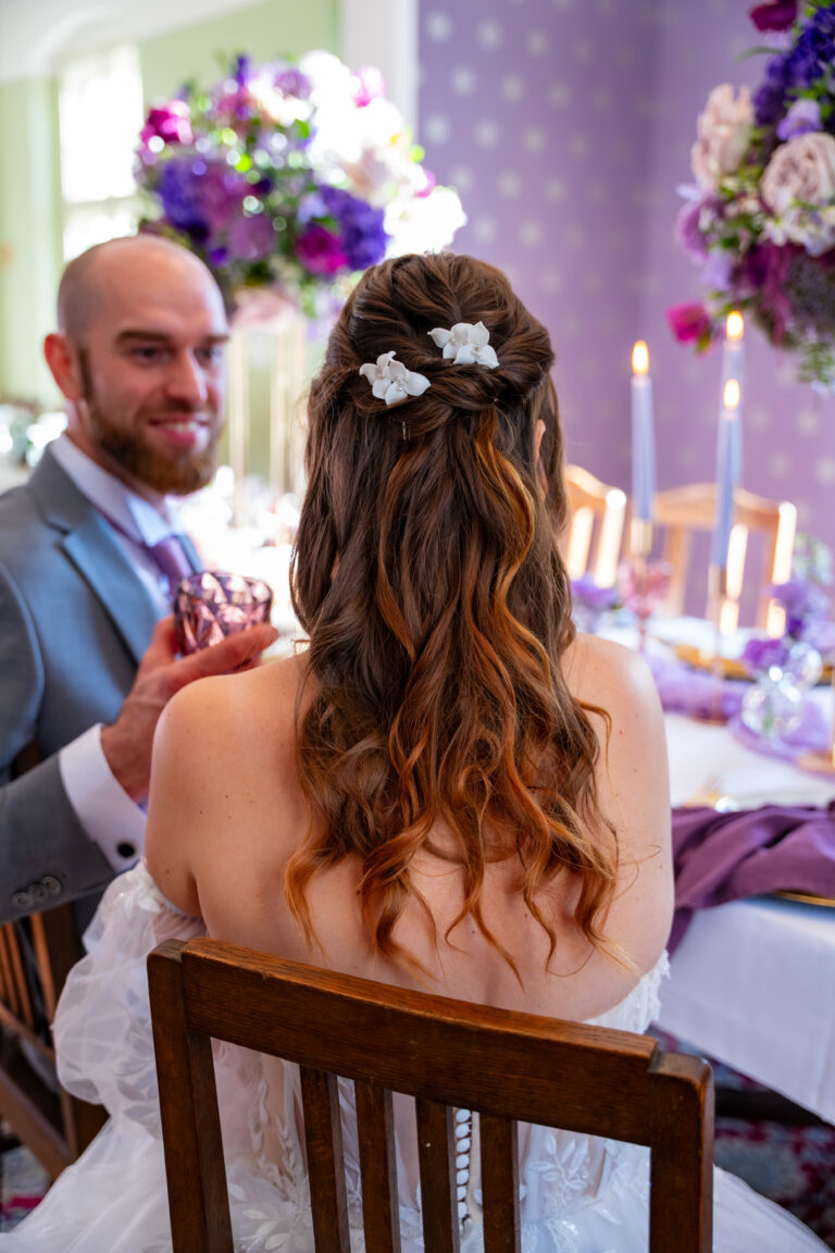 Braut mit gelocktem Haar und Blumenspangen sitzt an einem dekorierten Tisch mit wunderschöner Hochzeitsdeko neben einem lächelnden Bräutigam bei einem Hochzeitsempfang.