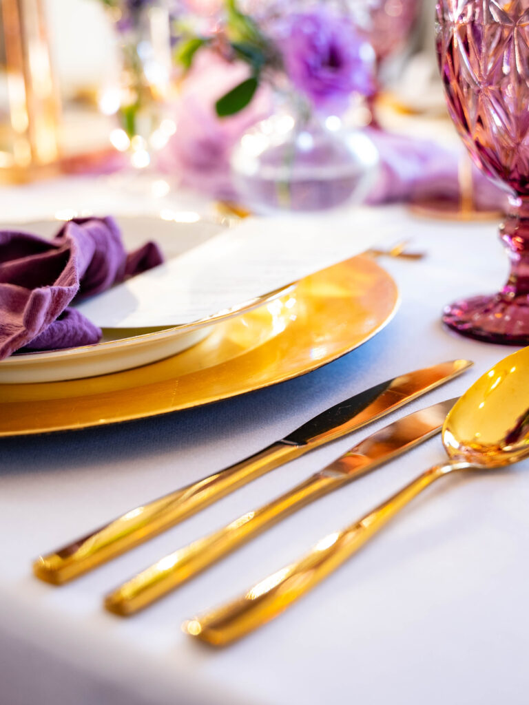 Elegant gedeckter Tisch mit goldenem Besteck, goldenem Platzteller, lila Serviette und rosa Glas auf weißer Tischdecke - die perfekte Hochzeitsdeko für Feiern in Brandenburg.