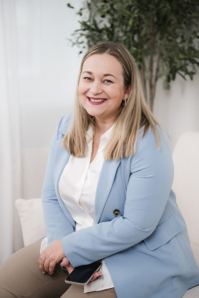Lächelnde Frau mit langen blonden Haaren in einem hellblauen Blazer sitzt in einem Haus mit einer Pflanze im Hintergrund, perfekt für alle, die eine Freie Traurednerin in Berlin oder Brandenburg suchen.