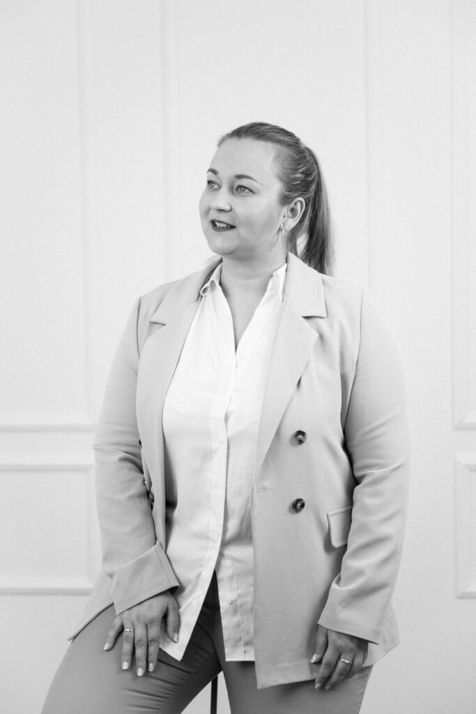 Eine Frau in Blazer und Bluse lächelt und schaut zur Seite in einer hellen, minimalistischen Umgebung - die perfekte Inspiration für eine Freie Traurednerin in Brandenburg.