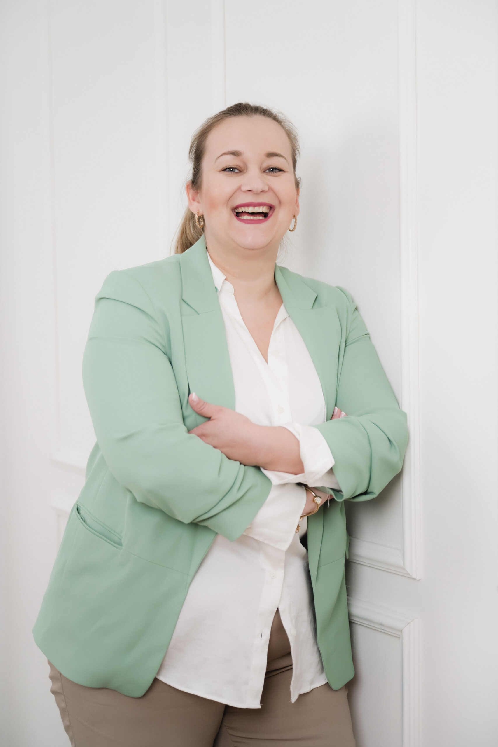 Freie Traurednerin aus Berlin in mintgrünem Blazer und weißem Shirt, lächelnd mit verschränkten Armen vor weißer Wand.