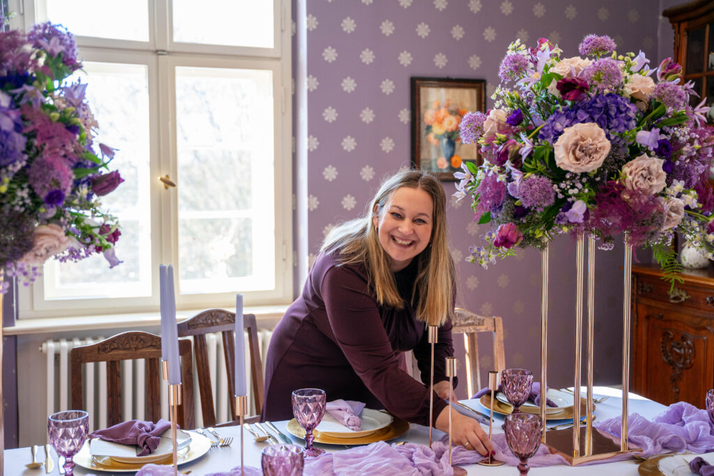 Eine lächelnde Frau deckt einen mit lila Blumen und Kerzen geschmückten Tisch in einem hell erleuchteten, eleganten Raum - perfekt für eine Freie Traurednerin in Berlin oder Brandenburg.