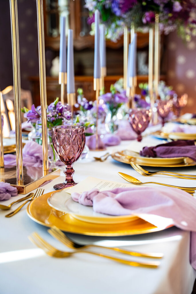 Elegant gedeckter Tisch mit goldenen Tellern, lila Servietten, Kristallgläsern und hohen Lavendelkerzen - die perfekte Hochzeitsdeko für Ihre Feier in Berlin.