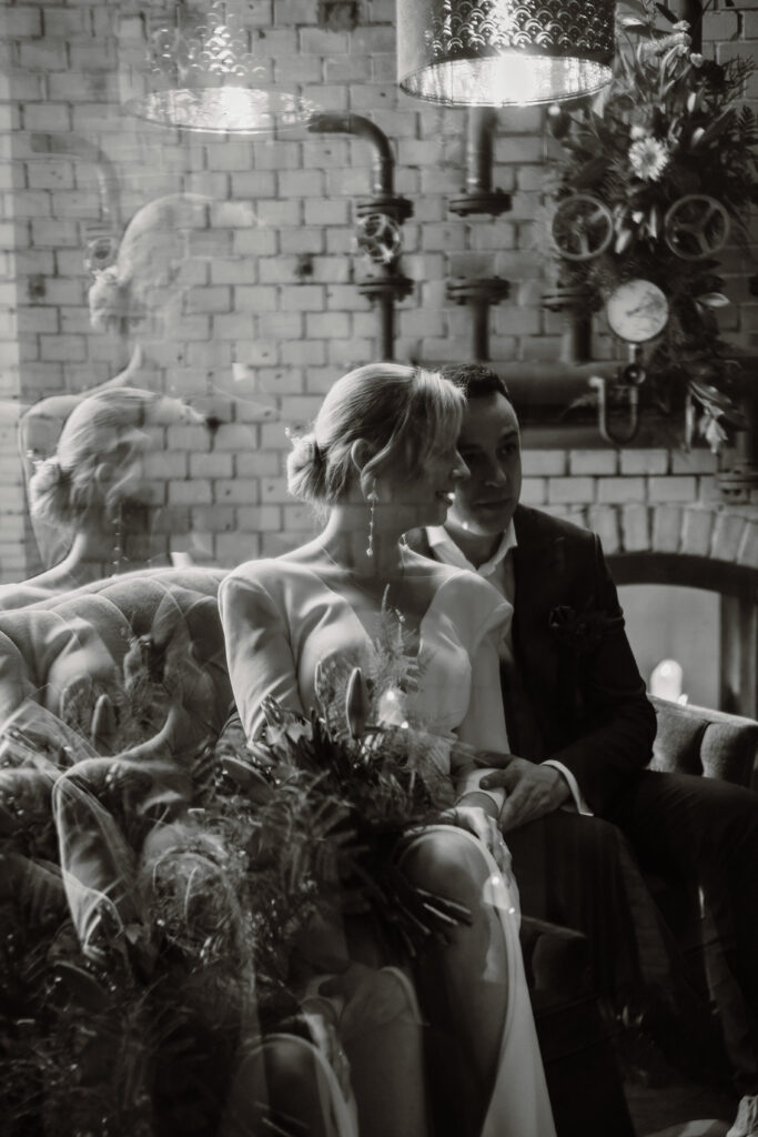 Braut und Bräutigam sitzen eng beieinander auf einer Couch, die sich in einem Glas spiegelt, in einem stimmungsvollen Raum im Industriestil - die perfekte Inspiration für alle Berliner Hochzeitsplaner, die auf der Suche nach einzigartigen Hochzeitsmomenten sind.