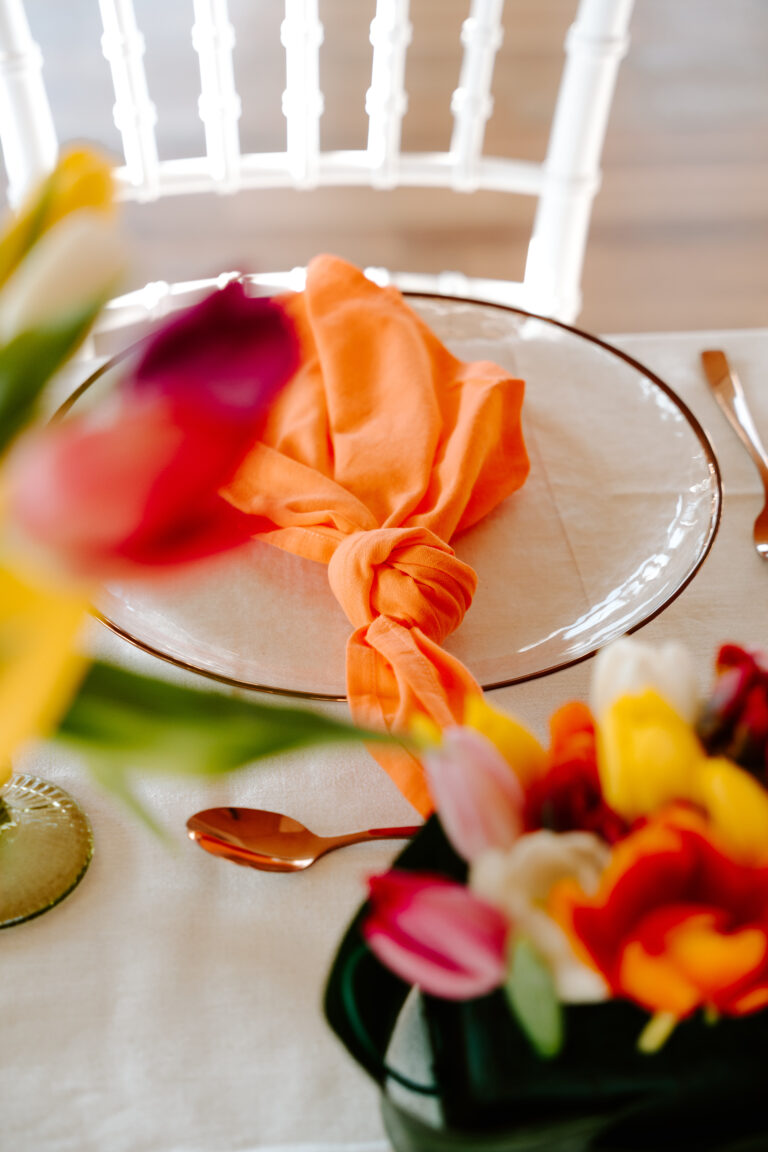 Ein gedeckter Tisch mit einer orangefarbenen Serviette, die auf einem Teller verknotet ist, und bunten Blumen im Vordergrund - die perfekte Inspiration für die Hochzeitsdeko von Ihrem Brandenburger Hochzeitsplaner.
