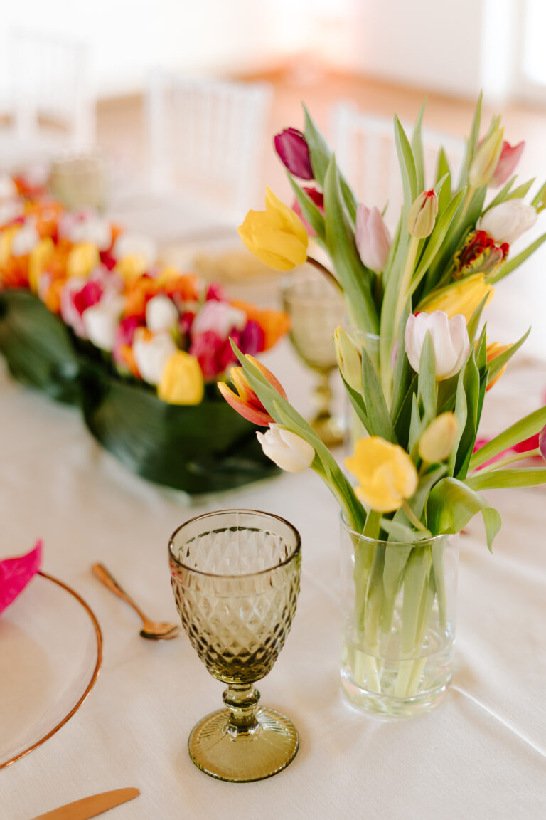 Eine Glasvase mit bunten Tulpen auf einem gedeckten Esstisch mit goldenem Geschirr und einem grünen Kelch - perfekt für eine elegante Hochzeitsdeko in Berlin.