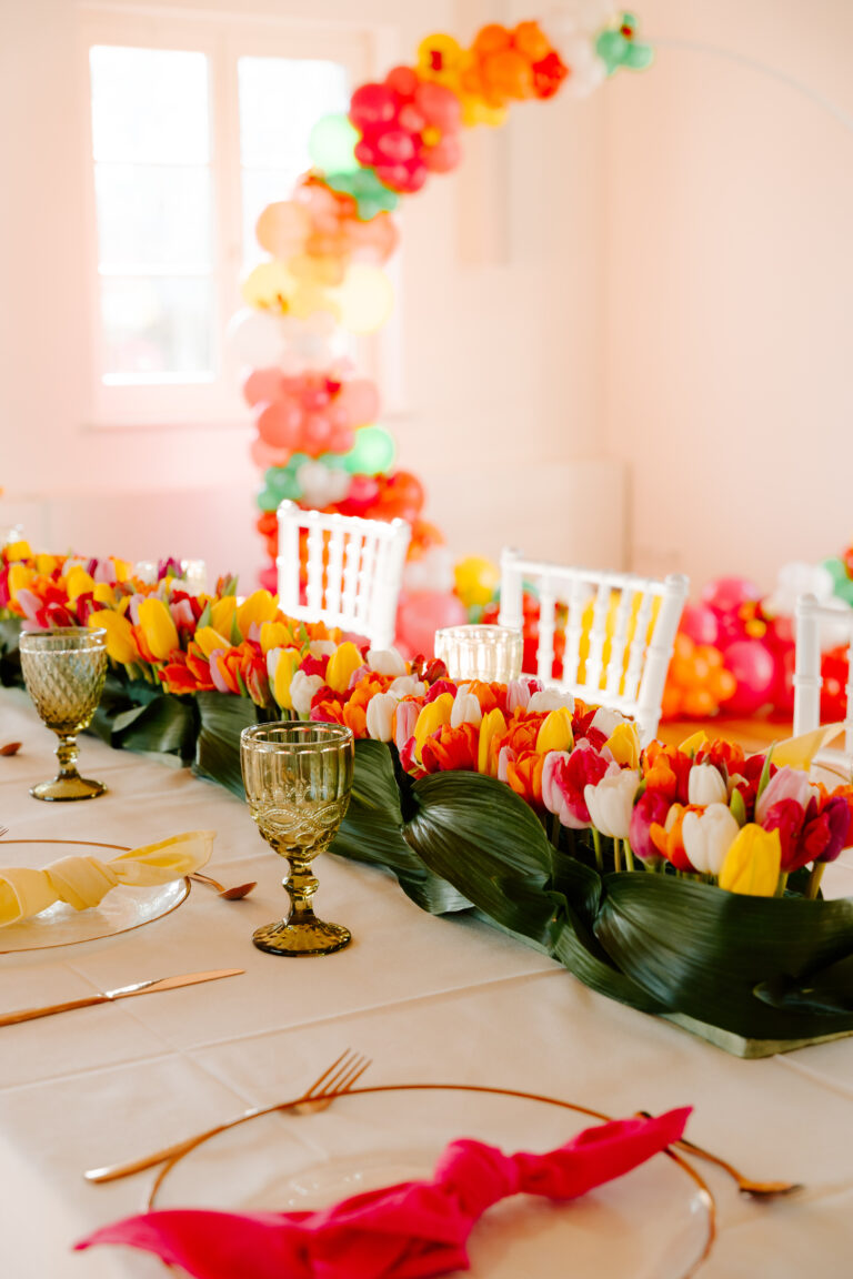 Elegant gedeckter Tisch mit farbenfrohem Tulpen-Mittelstück, goldenen Utensilien und weißen Stühlen; atemberaubender Ballonbogen im Hintergrund - die perfekte Hochzeitsdeko-Inspiration für Feiern in Berlin oder Brandenburg.