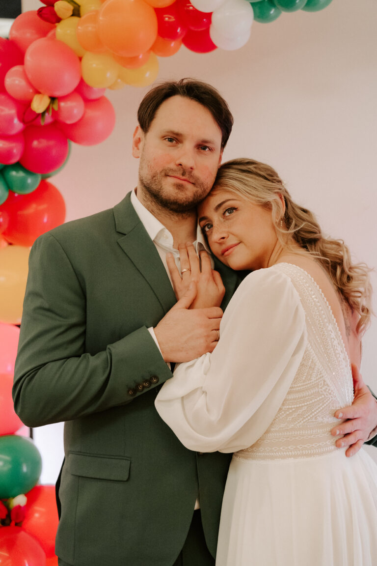 Ein Paar in Hochzeitskleidung umarmt sich unter einem farbenfrohen Luftballonbogen - eine bezaubernde Hochzeitsdeko-Szene in Berlin - und blickt sanft in die Kamera.
