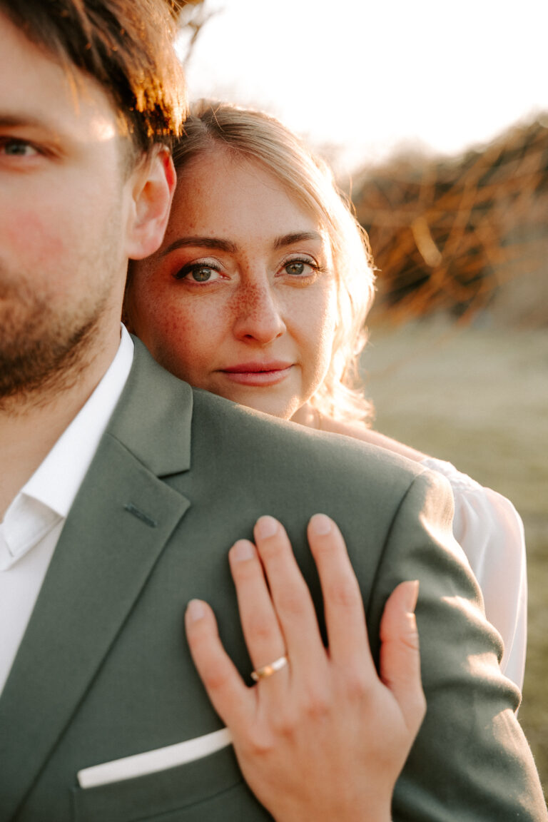 Eine Frau umarmt einen Mann von hinten, ihre Hand ruht auf seiner Schulter, beide im sanften Sonnenlicht - ein intimer Moment, perfekt für einen Hochzeitsplaner in Brandenburg.