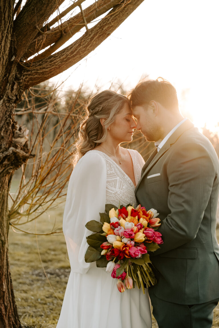 Braut und Bräutigam umarmen sich unter einem Baum bei Sonnenuntergang in Brandenburg, in der Hand einen bunten Tulpenstrauß, umgeben von eleganter Hochzeitsdeko.