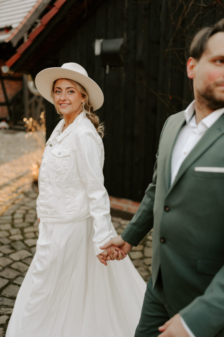 Eine Braut in weißem Kleid und Hut hält Händchen mit einem Bräutigam in grünem Anzug, der im Freien in Brandenburg spazieren geht, umgeben von einer eleganten Hochzeitsdeko.