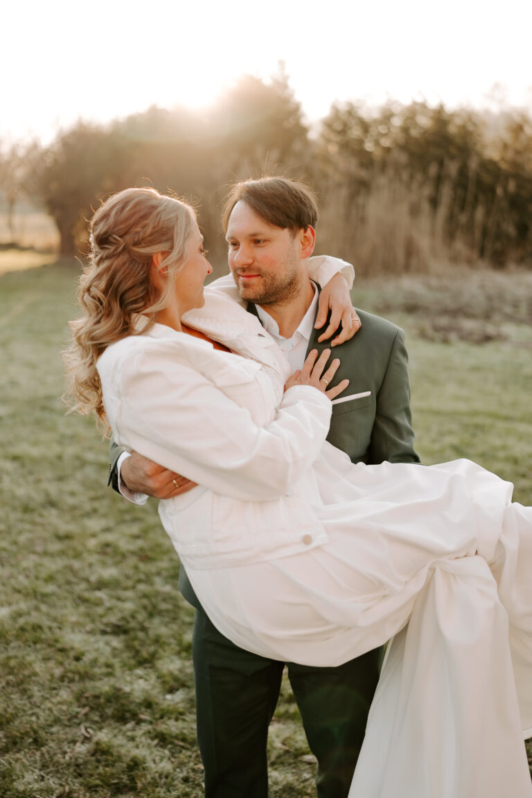 Ein Bräutigam trägt seine Braut bei Sonnenuntergang im Freien in Brandenburg, beide lächeln und schauen sich liebevoll an.