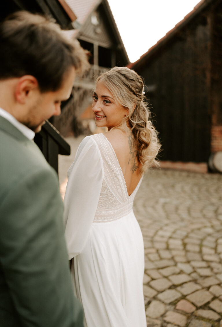 Eine Braut in einem weißen Kleid lächelt einem Mann im Anzug auf einem kopfsteingepflasterten Weg im Freien zu, umgeben von einer eleganten Hochzeitsdeko, die den Charme der romantischen Hochzeitsszene Berlins einfängt.