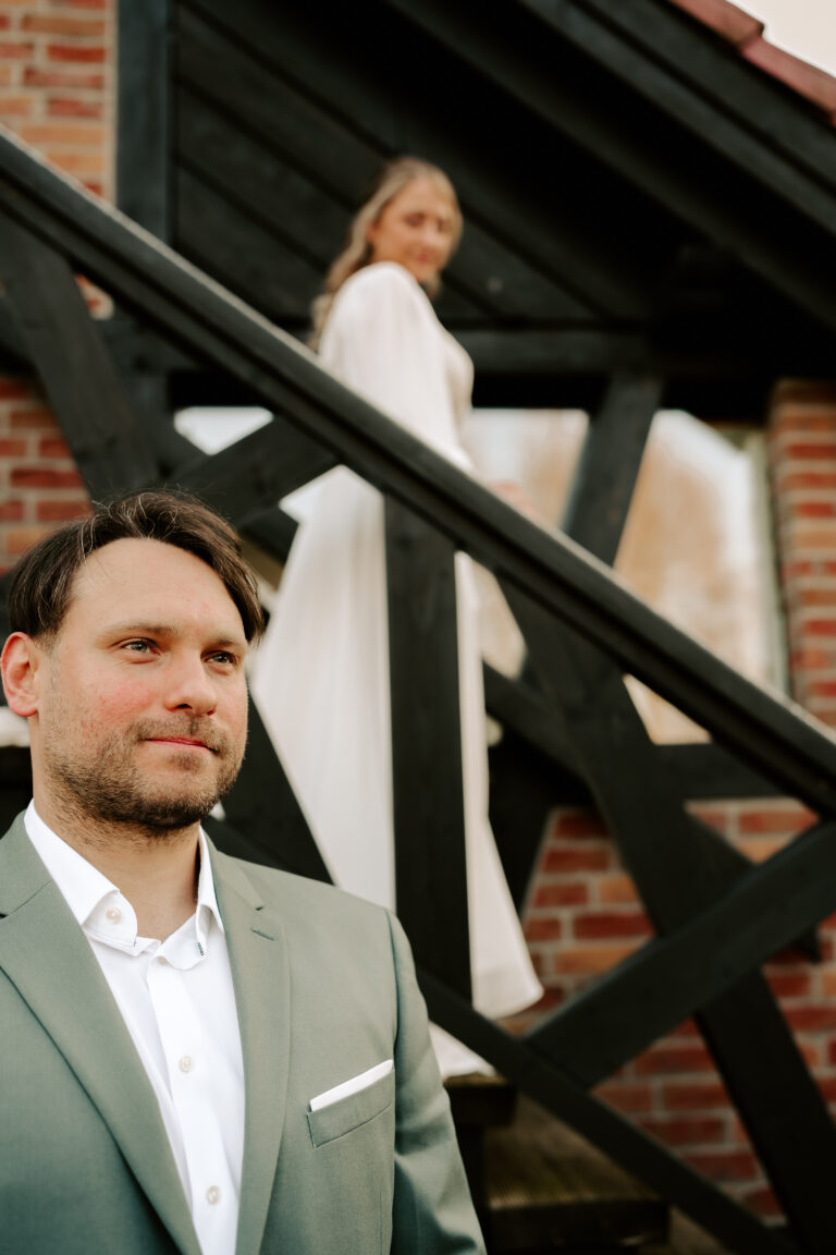 Ein Mann im Anzug steht im Mittelpunkt, eine Frau im weißen Kleid steht unscharf auf einer Treppe hinter ihm, vor der reizvollen Kulisse von Berlin.