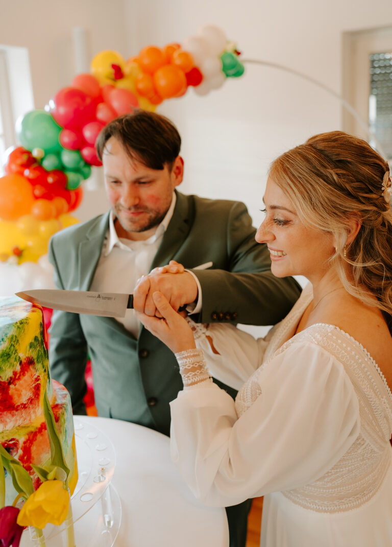 Braut und Bräutigam lächelnd beim Anschneiden einer bunten Torte, mit Luftballons im Hintergrund, auf ihrer Berliner Feier.