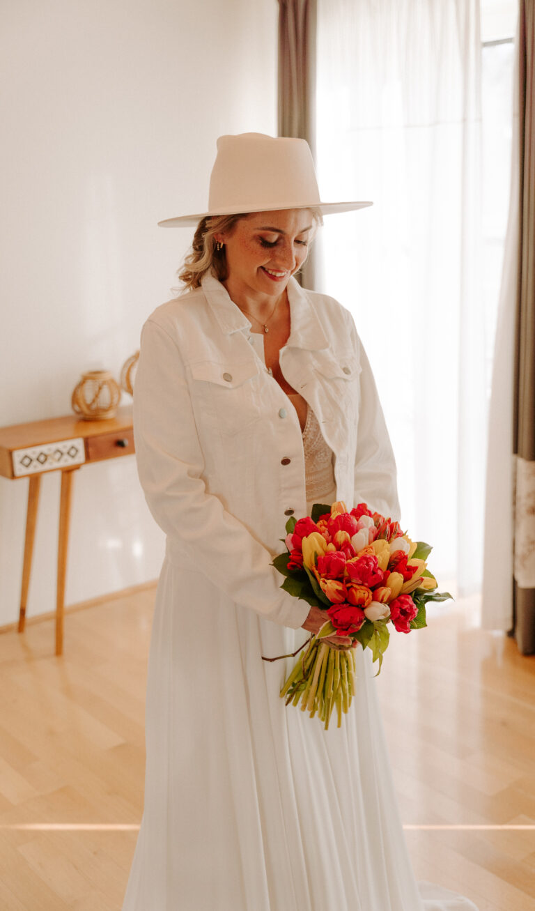 Eine lächelnde Braut in weißem Kleid und Hut hält einen bunten Blumenstrauß in der Hand, umgeben von eleganter, von Berlin und Brandenburg inspirierter Hochzeitsdeko.